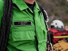 Load image into Gallery viewer, Person wearing a green uniform with a name tag reading 'J. Wilson Rescue' in an outdoor setting.