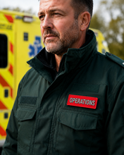 Load image into Gallery viewer, A close-up of a man wearing a dark green tactical jacket with a red 'OPERATIONS' patch on the chest, standing in front of a yellow ambulance.