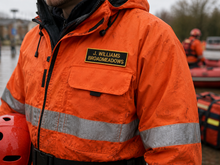 Load image into Gallery viewer, Person wearing an orange jacket with a name tag and reflective stripes, standing outdoors.

