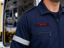 Load image into Gallery viewer, Person wearing a navy blue uniform with a name tag labeled 'Wilson' in front of an ambulance.
