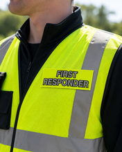 Load image into Gallery viewer, A close-up of a person wearing a high-visibility neon yellow safety vest with reflective gray stripes and a yellow patch that reads 'FIRST RESPONDER' in black letters. The vest is worn over a black zip-up jacket outdoors.
